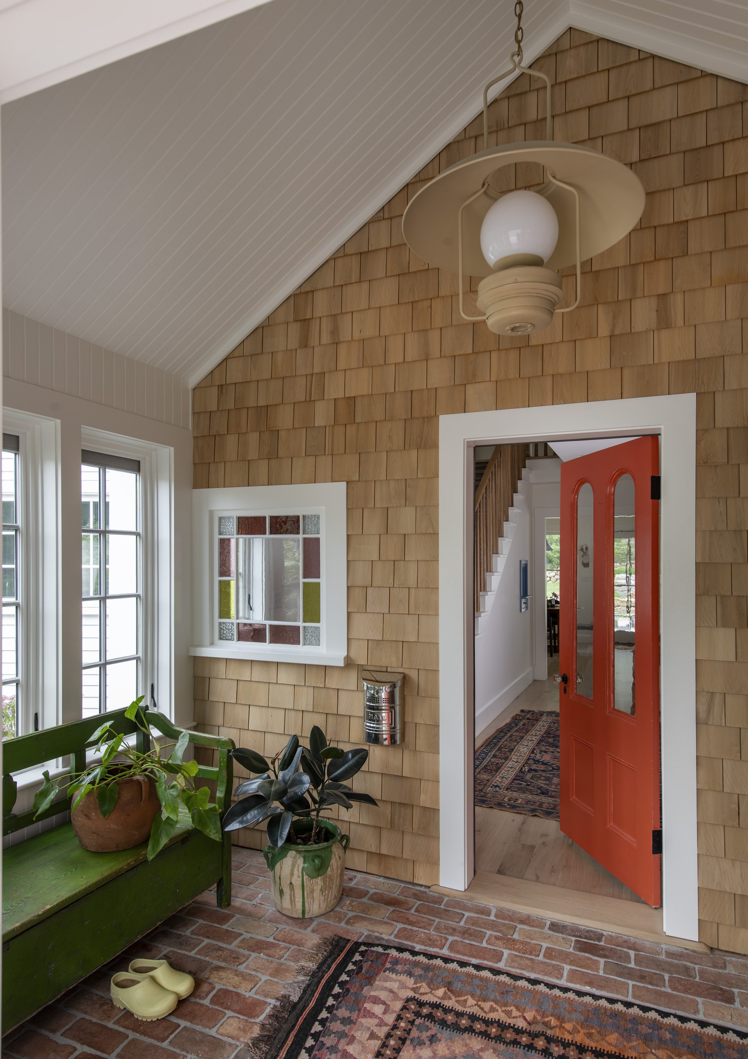 Interior view of a room with a ceiling-mounted vintage lamp, brick floor, and wood-shingle wall; open front door reveals stairs inside, potted plants by a window, and a colorful rug.