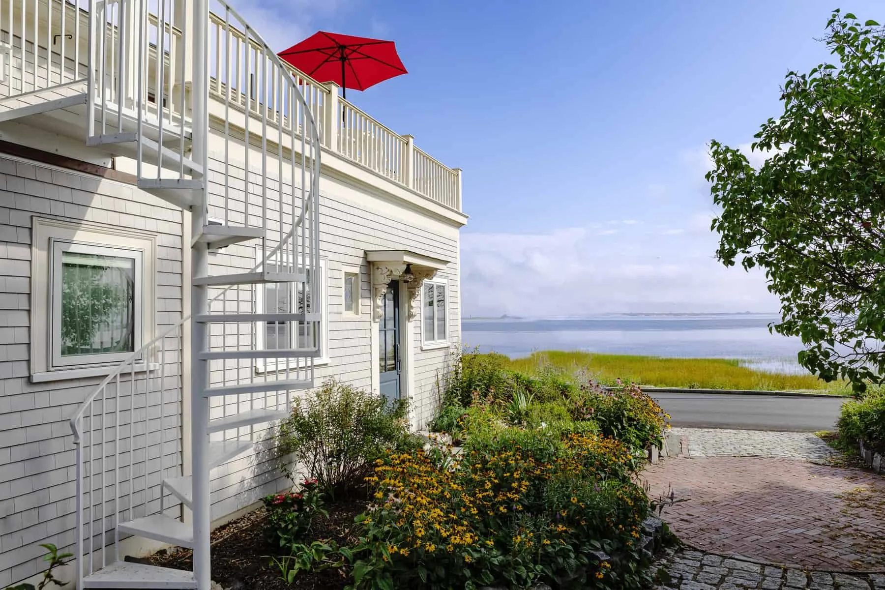 A white house with vinyl siding and a blue door, featuring an exterior spiral staircase leading to a balcony with a red patio umbrella. The house is surrounded by colorful flowers and greenery, with a view of the water and a clear blue sky in the bac
