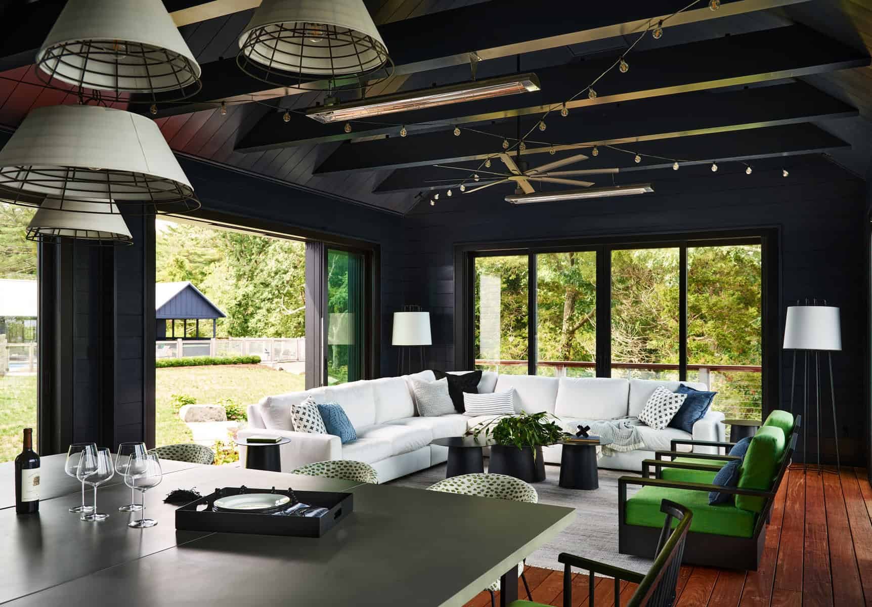 Modern indoor living room with large windows, white sectional sofa, green chairs, black coffee tables, and decorative pillows. Ceiling features black beams, large industrial-style pendant lights, and string lights. Exterior view shows trees, a small 