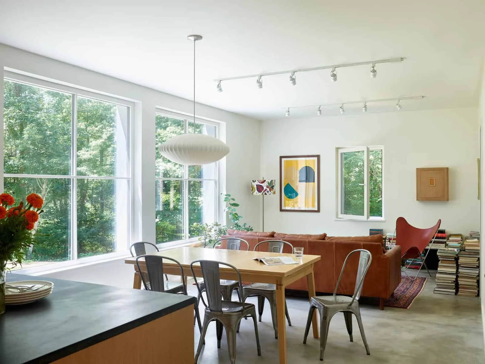 Bright living room with large windows, white walls, modern art, a brown sectional sofa, colorful chairs, houseplants, stacks of books, and decorative lighting.