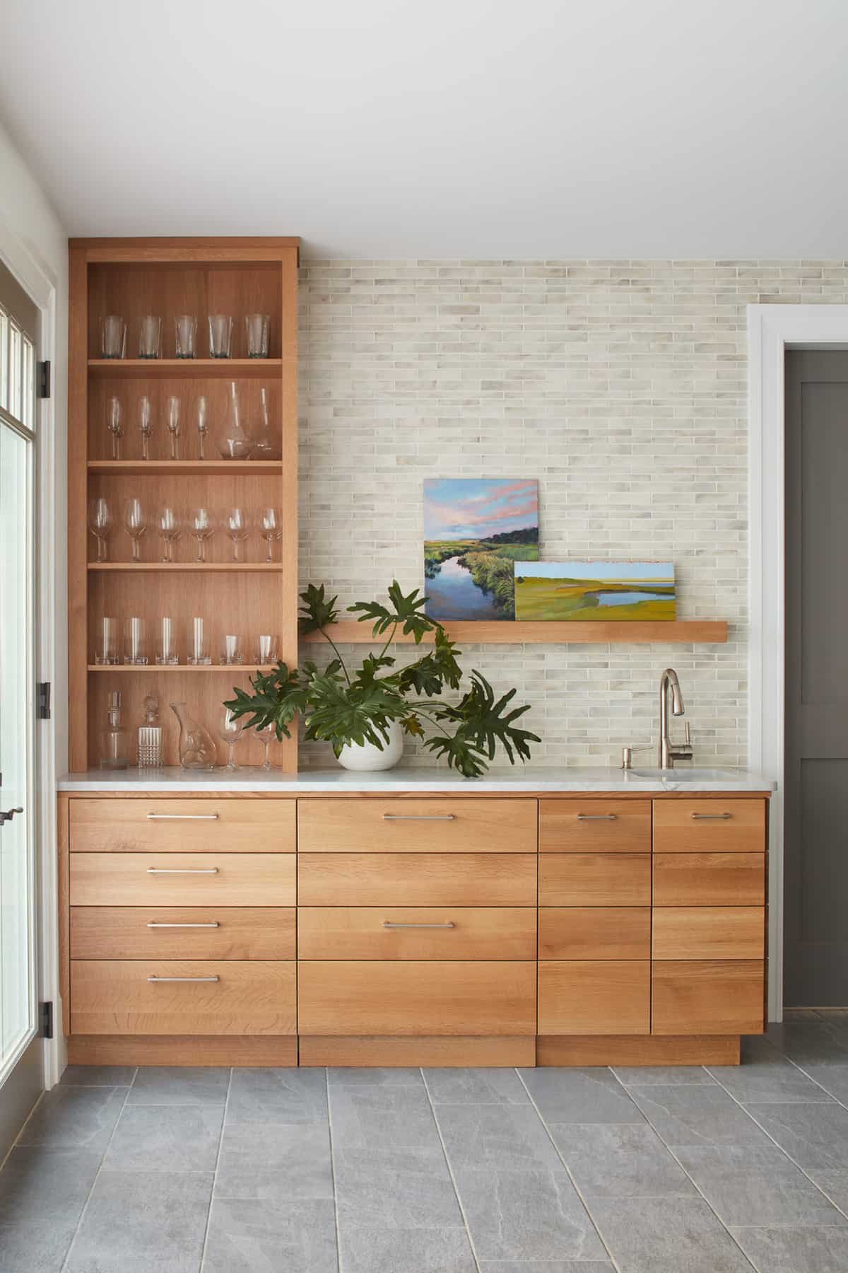 Modern kitchen with light wood cabinets, a white marble countertop, glassware on open shelves, a potted plant, and landscape paintings on a textured brick wall.