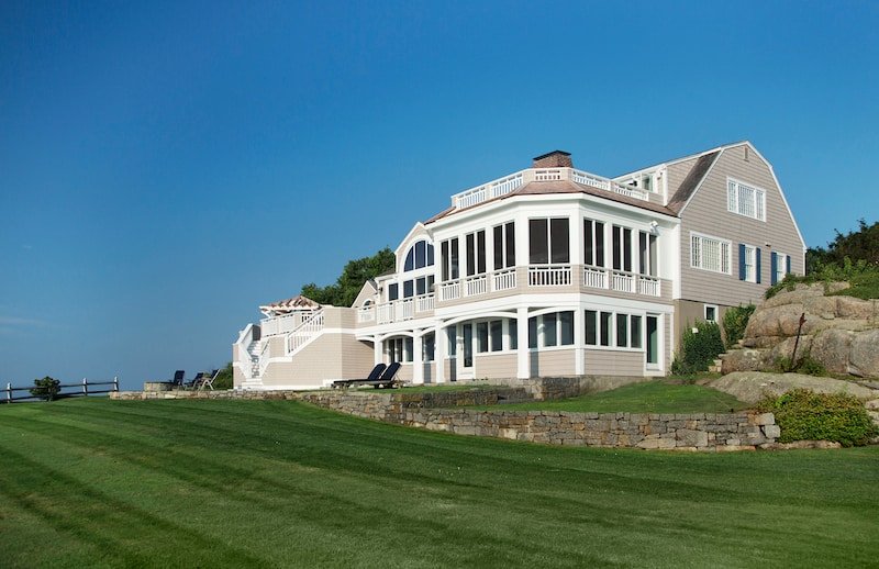Large coastal house with multiple floors and a wraparound balcony, set on a grassy hill with a stone retaining wall, under a clear blue sky.