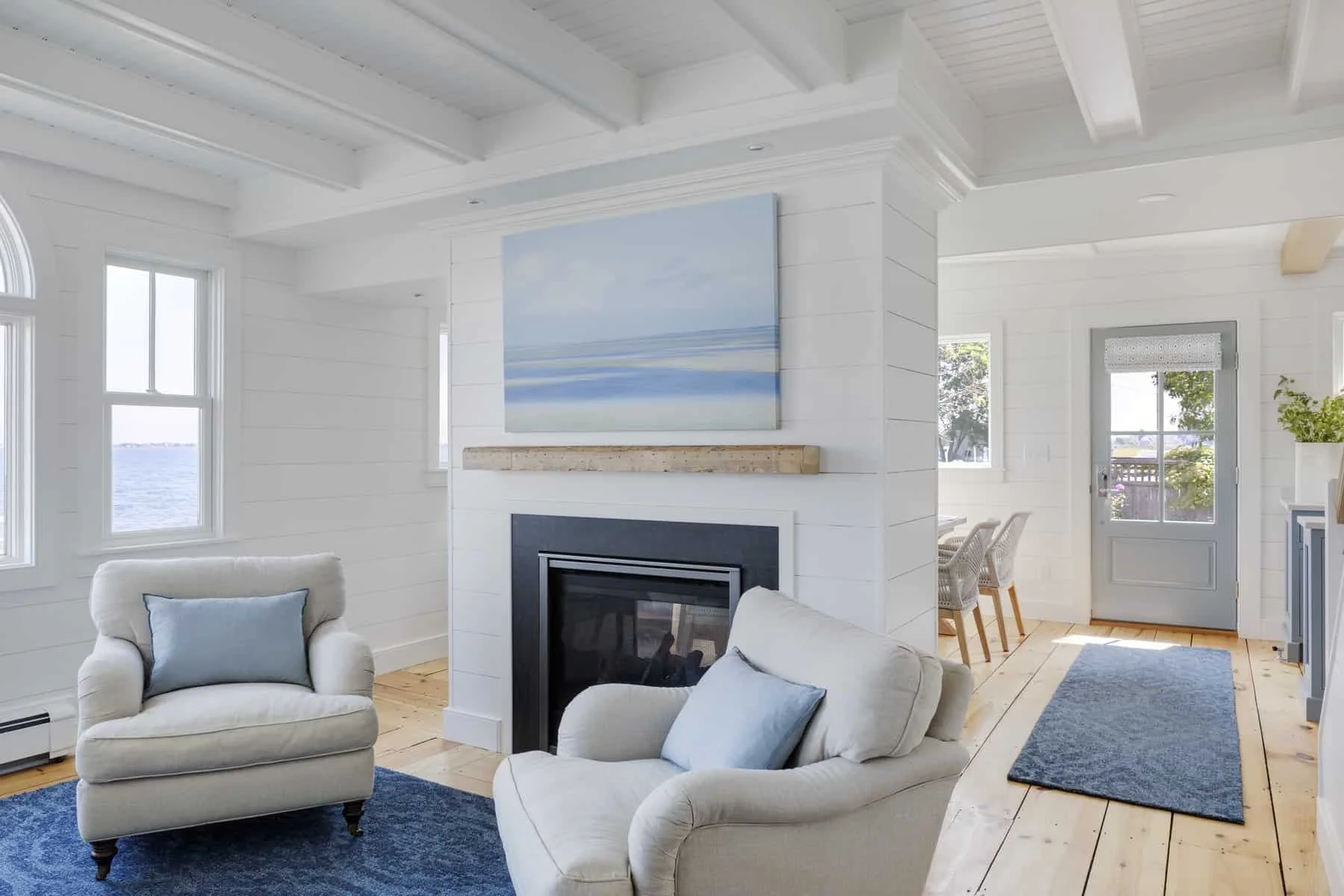 Bright living room with white walls, featuring a fireplace, framed ocean view through the windows, and a cozy seating area with white armchairs and blue accents.