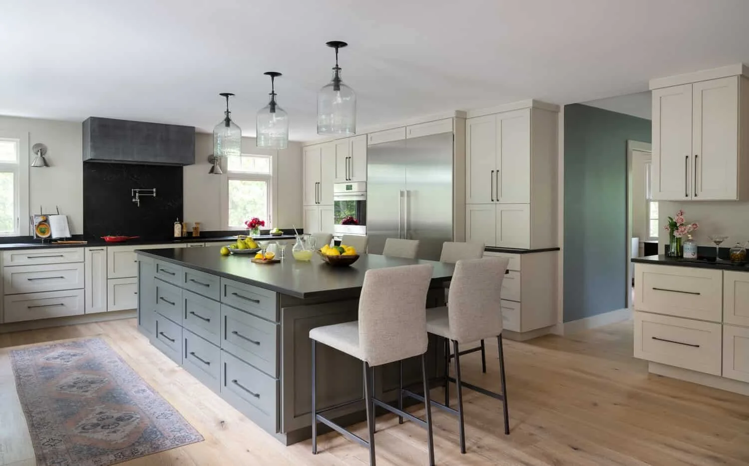 Modern kitchen with white cabinets, gray island with drawers, three glass pendant lights, black countertops, and stainless steel appliances. There are flowers and fruit on the counters and island, and a rug on the wooden floor.