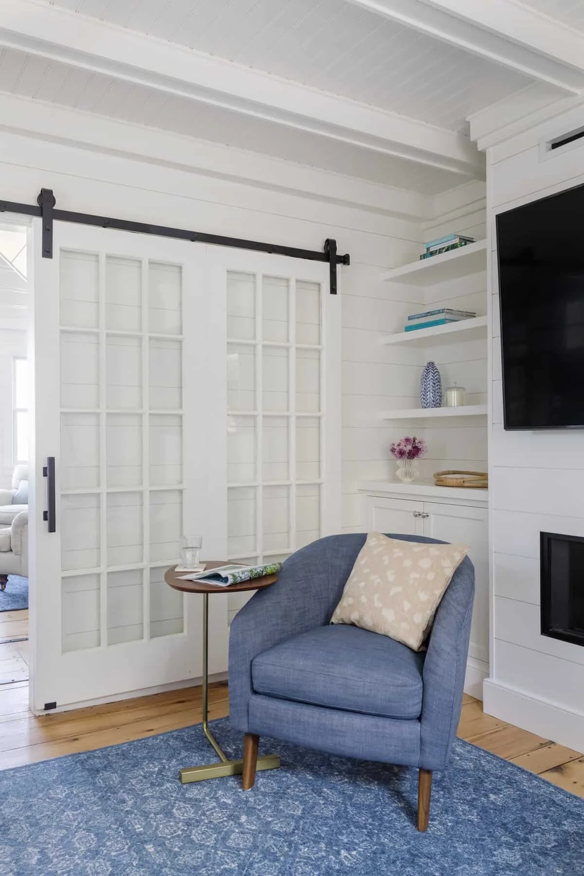 A cozy living room corner with a blue armchair, beige patterned pillow, small side table with a glass of water and open magazine, white shelving with books and decorative items, white sliding door with black hardware, and a mounted flat-screen TV.