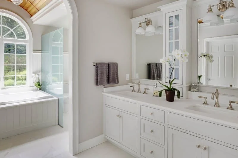 Bright bathroom with white cabinetry, dual sinks, large mirror, and a window with a view of greenery.