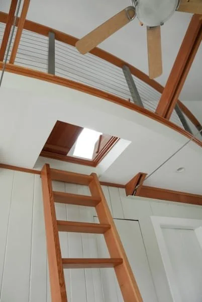 Interior view of a room with a wooden attic ladder extending to a loft with a ceiling fan, wooden beams, and a skylight.