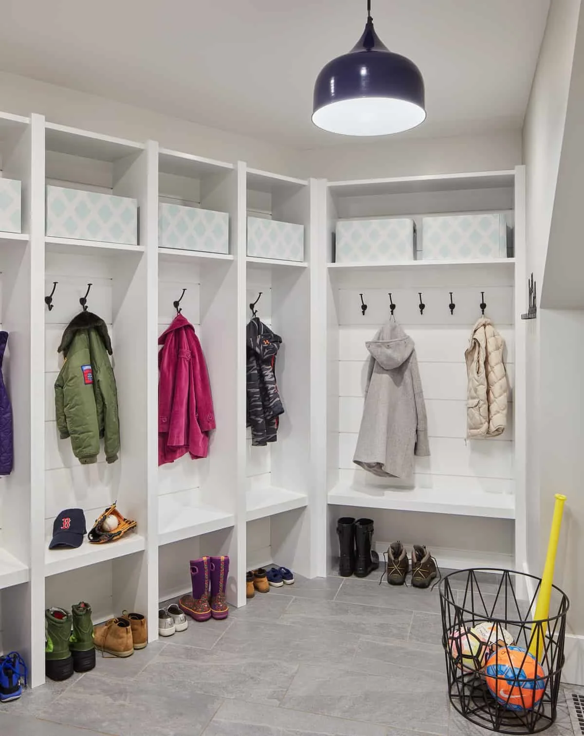 A bright mudroom with white built-in cubbies and hooks, holding jackets and coats. Shoes and boots are organized below, and a black wire basket with balls is on the floor. A black ceiling light hangs overhead.