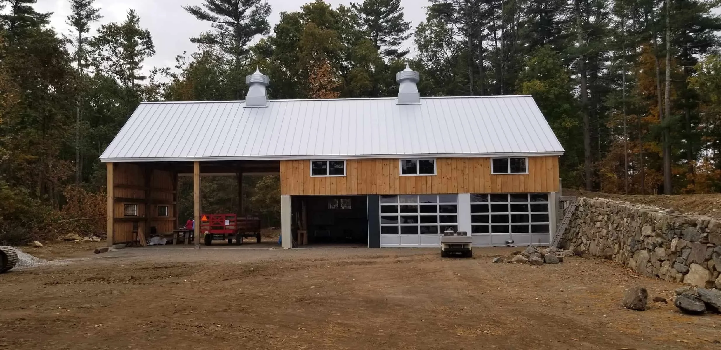 Under construction house with a metal roof, wooden siding, and a garage door, surrounded by trees and dirt ground.