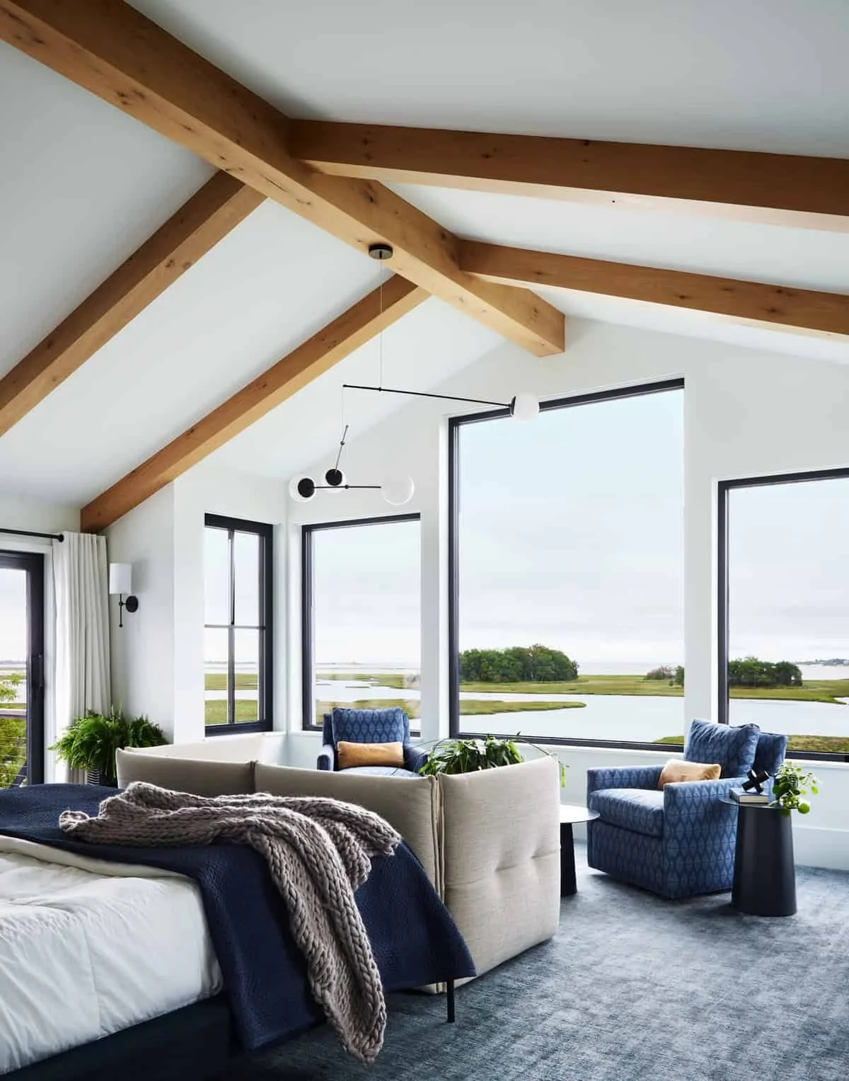 Living room with large windows, white walls, wooden ceiling beams, modern lighting, and blue and beige furniture overlooking a body of water and green landscape.