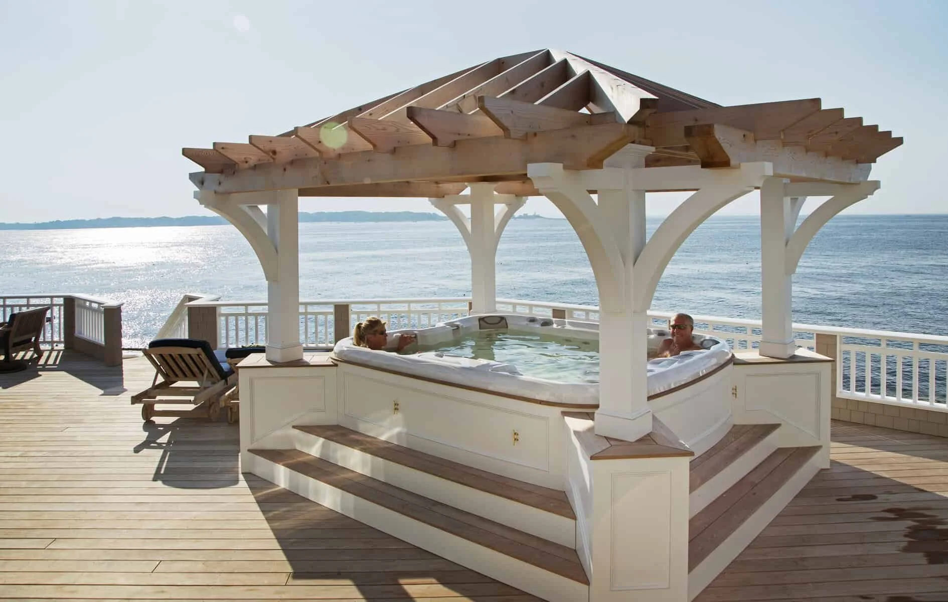 Two people relaxing in a hot tub on a deck overlooking the ocean, with a wooden gazebo structure overhead and picnic chairs nearby.