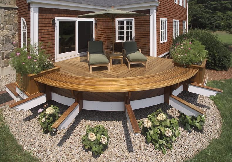 Wooden backyard deck with two lounge chairs, a small table, green cushions, an umbrella, built-in flower boxes, and surrounding plants and gravel.