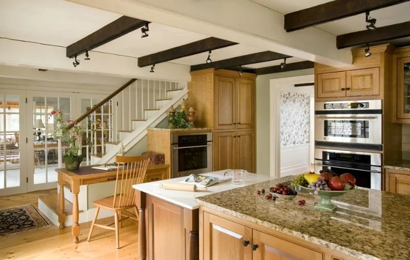 Kitchen with wooden cabinets, granite countertop island, built-in oven, and a staircase in the background. Fruit bowl and potted plant on the kitchen table.