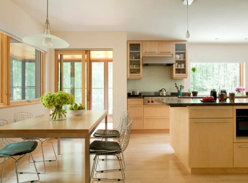 Bright, modern kitchen and dining area with large windows, wooden cabinets, and a central island. A dining table with a vase of green flowers and black chairs is visible.