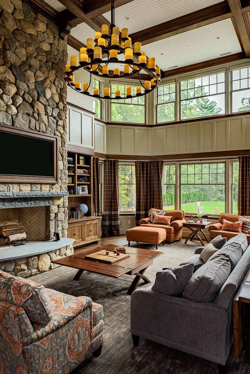 A cozy living room with large windows, a stone fireplace, a wooden coffee table, a gray sofa, floral armchair, and orange upholstered chairs, illuminated by a multi-tiered chandelier with yellow candle-like lights.