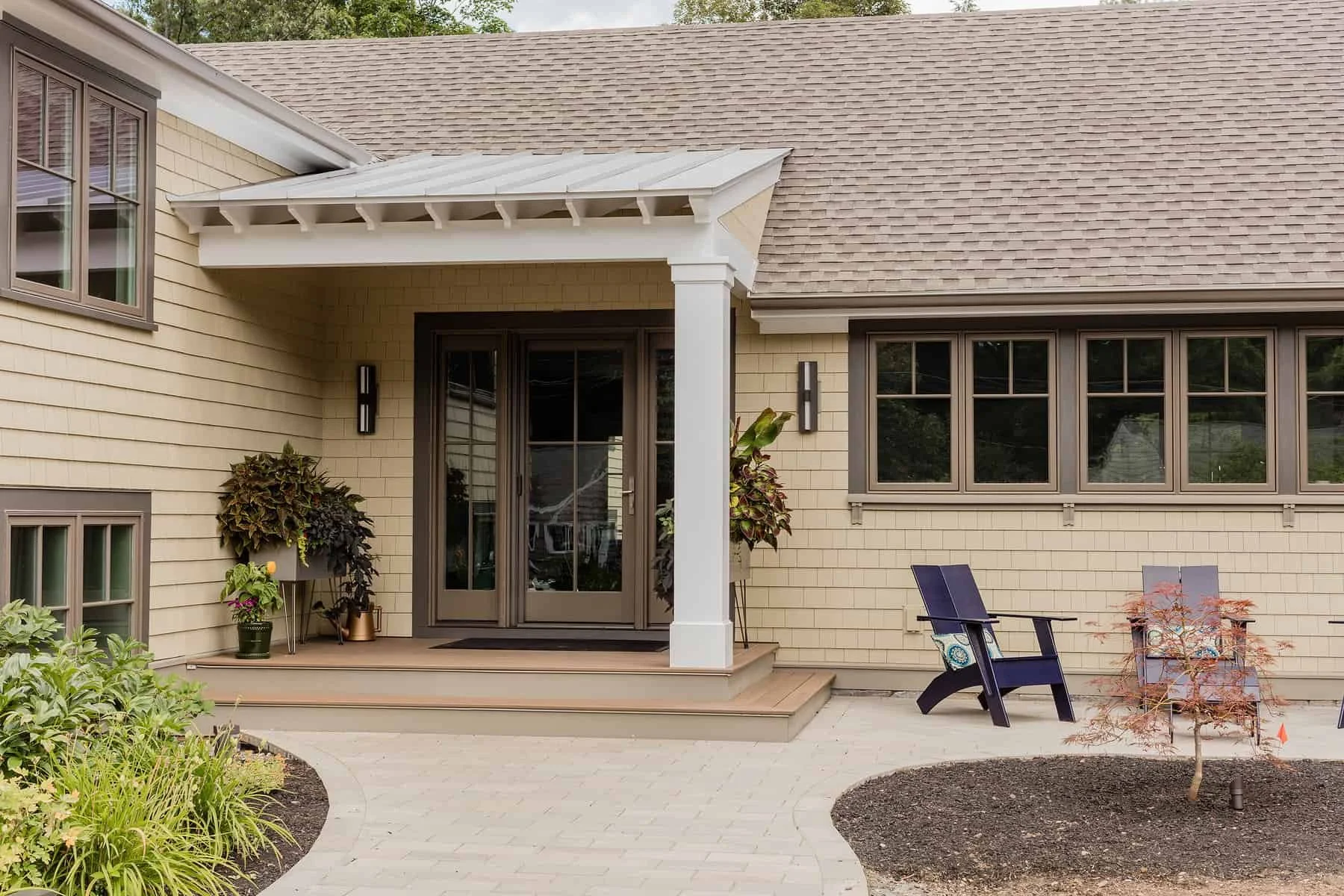 Backyard patio area with a small set of stairs leading up to a screened-in door. Decorated with potted plants, a blue chair, a gray chair, and a small tree with reddish leaves. The house has beige siding, multiple windows, and a gabled roof.