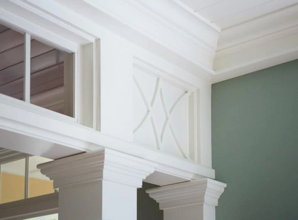 Close-up of a corner of a room showing white crown moulding, columns, a window with divided panes, and a wall painted in soft green.