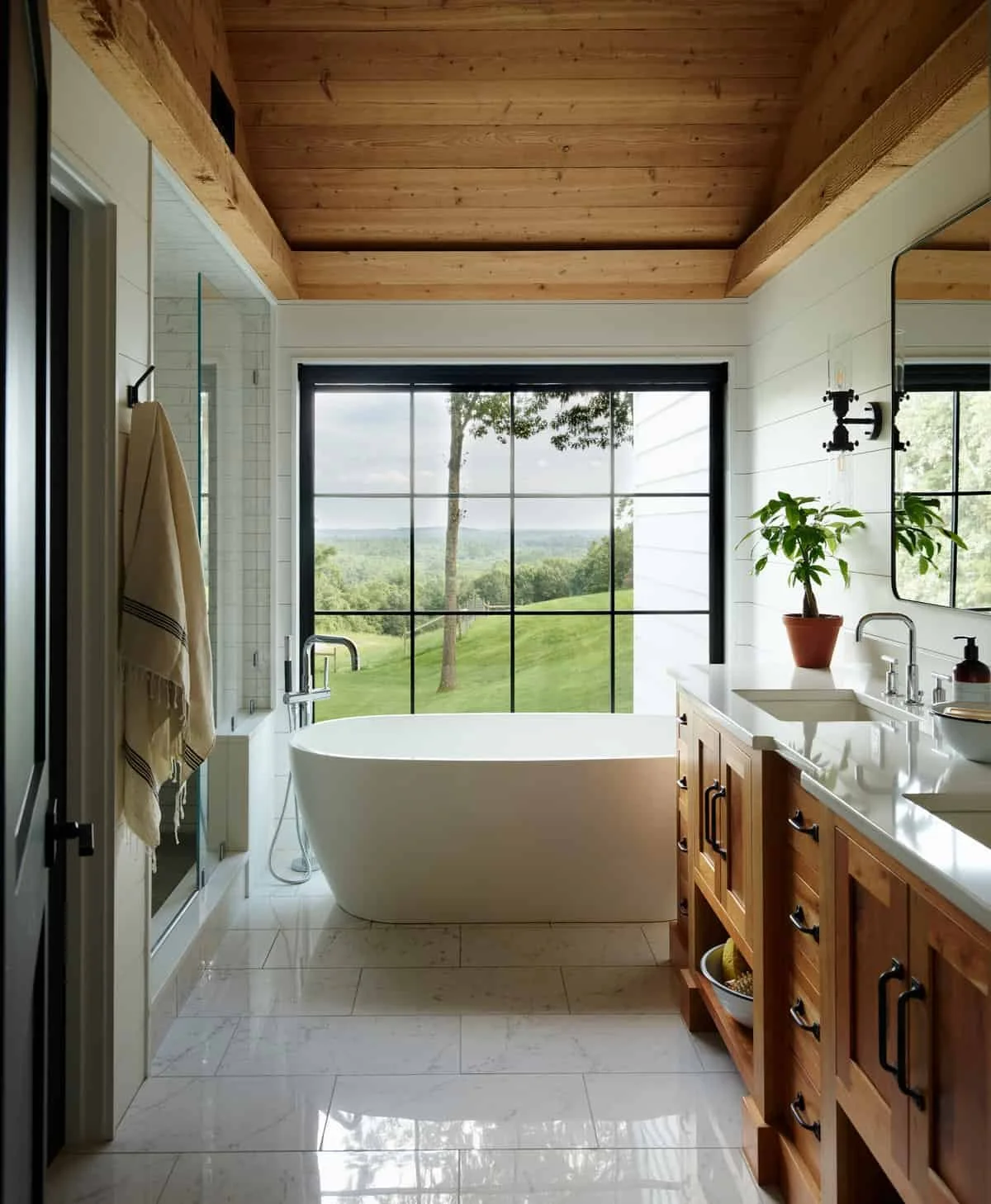 Modern bathroom with a large window overlooking a green landscape, featuring a freestanding bathtub, wooden cabinetry, potted plant, and a wood-paneled ceiling.