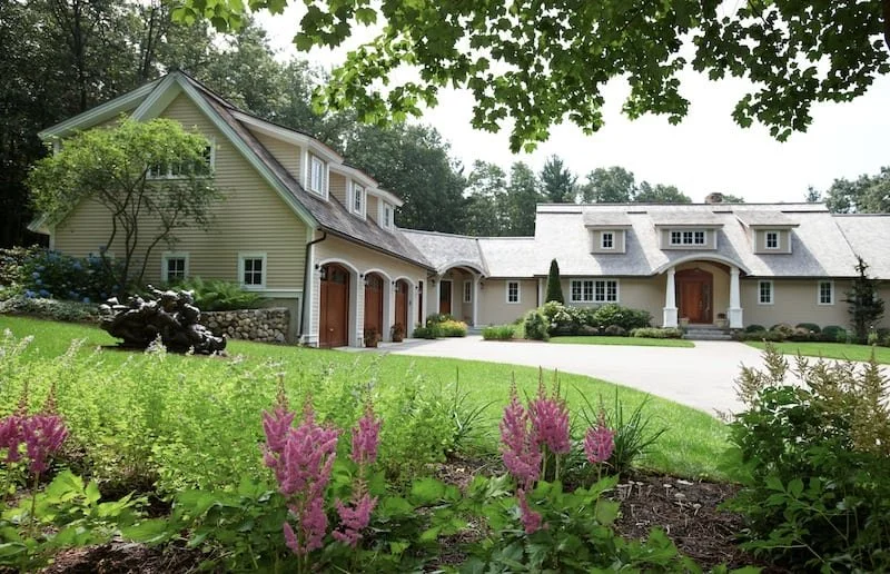Large house with multiple garage doors, set in a landscaped yard with pink flowers and green trees.