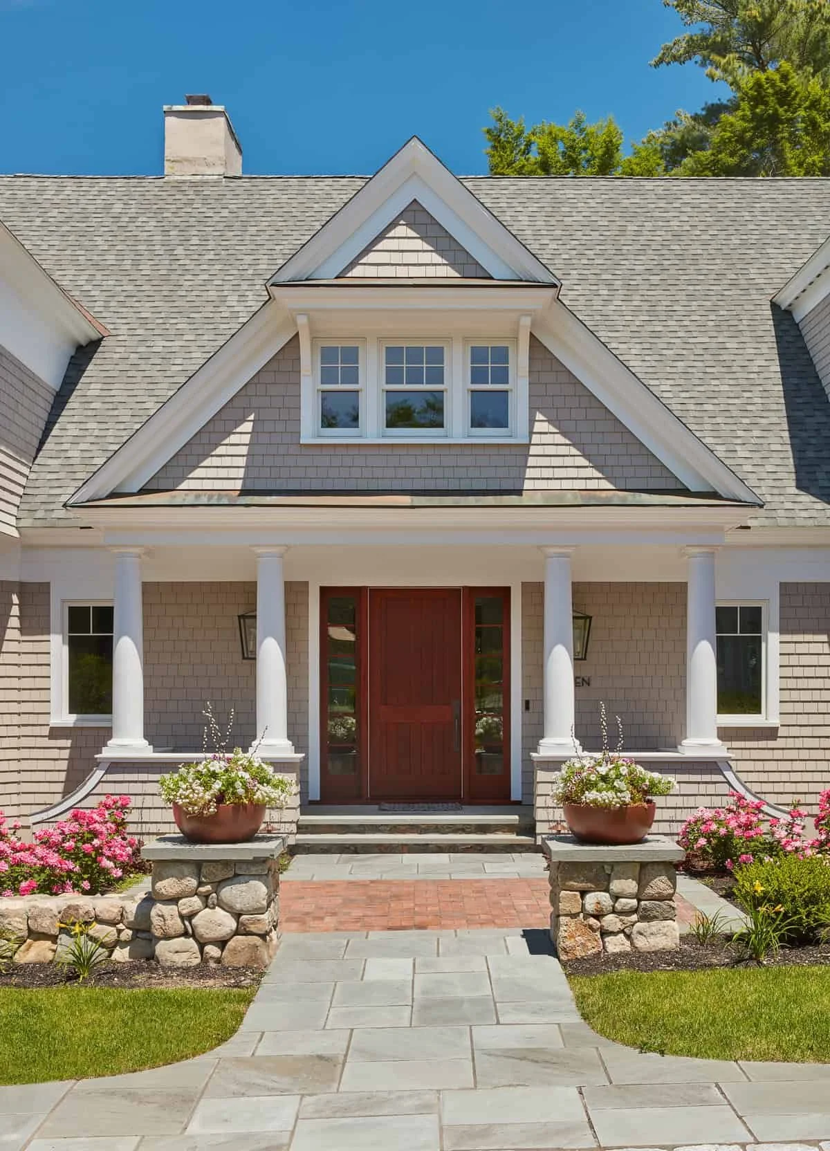 Front view of a house with gray shingle exterior, gray roof, white columns, and a red front door; landscaped yard with flower pots, pink and white flowers, and a stone pathway.