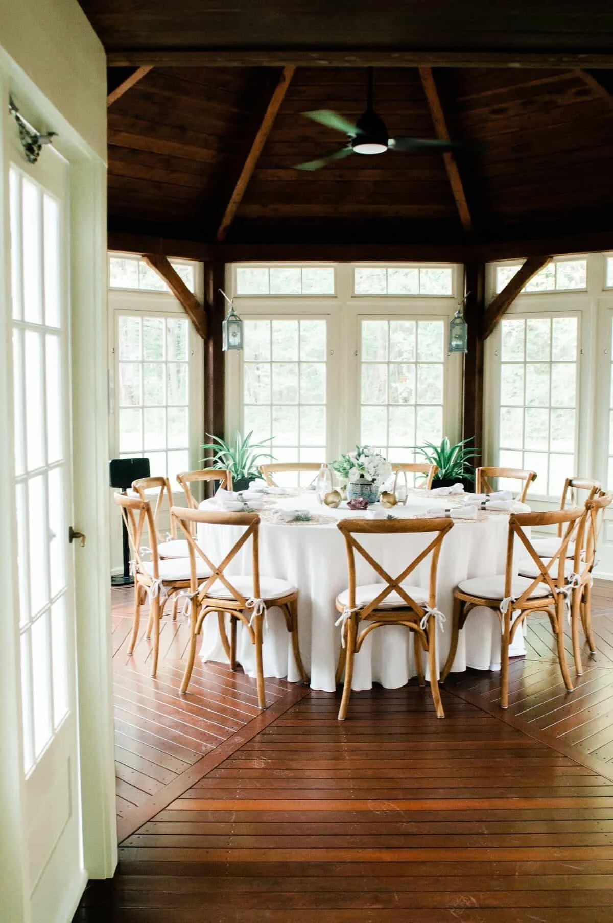 A round dining table set for a meal, surrounded by wooden chairs with white cushions, in a sunlit room with large windows, wooden floors, and a dark wooden ceiling fan.
