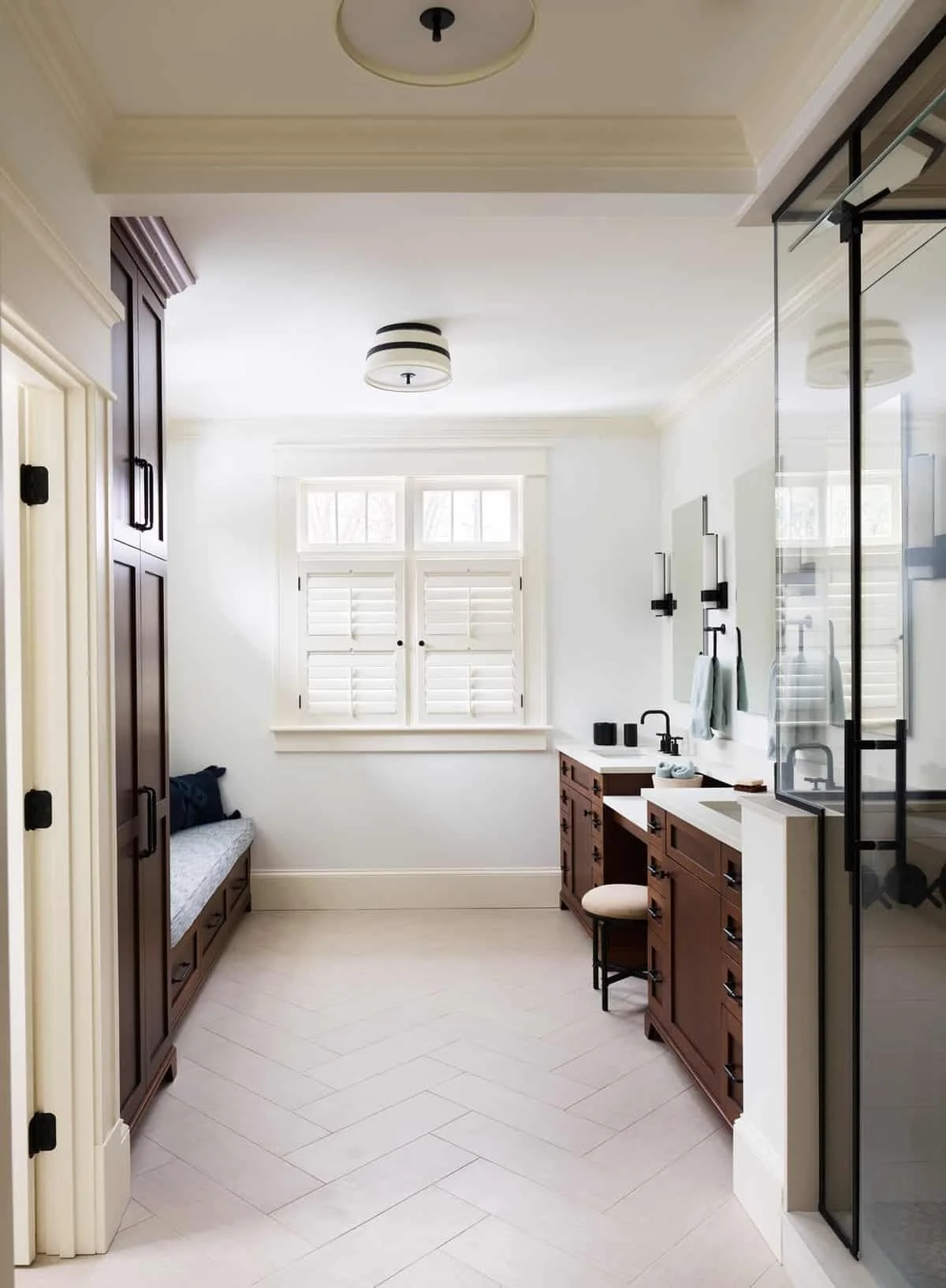 Bright bathroom with white walls, large window with shutters, dark wooden vanity, marble countertop, black fixtures, glass shower enclosure, built-in bench with black pillow, and minimalist decor.