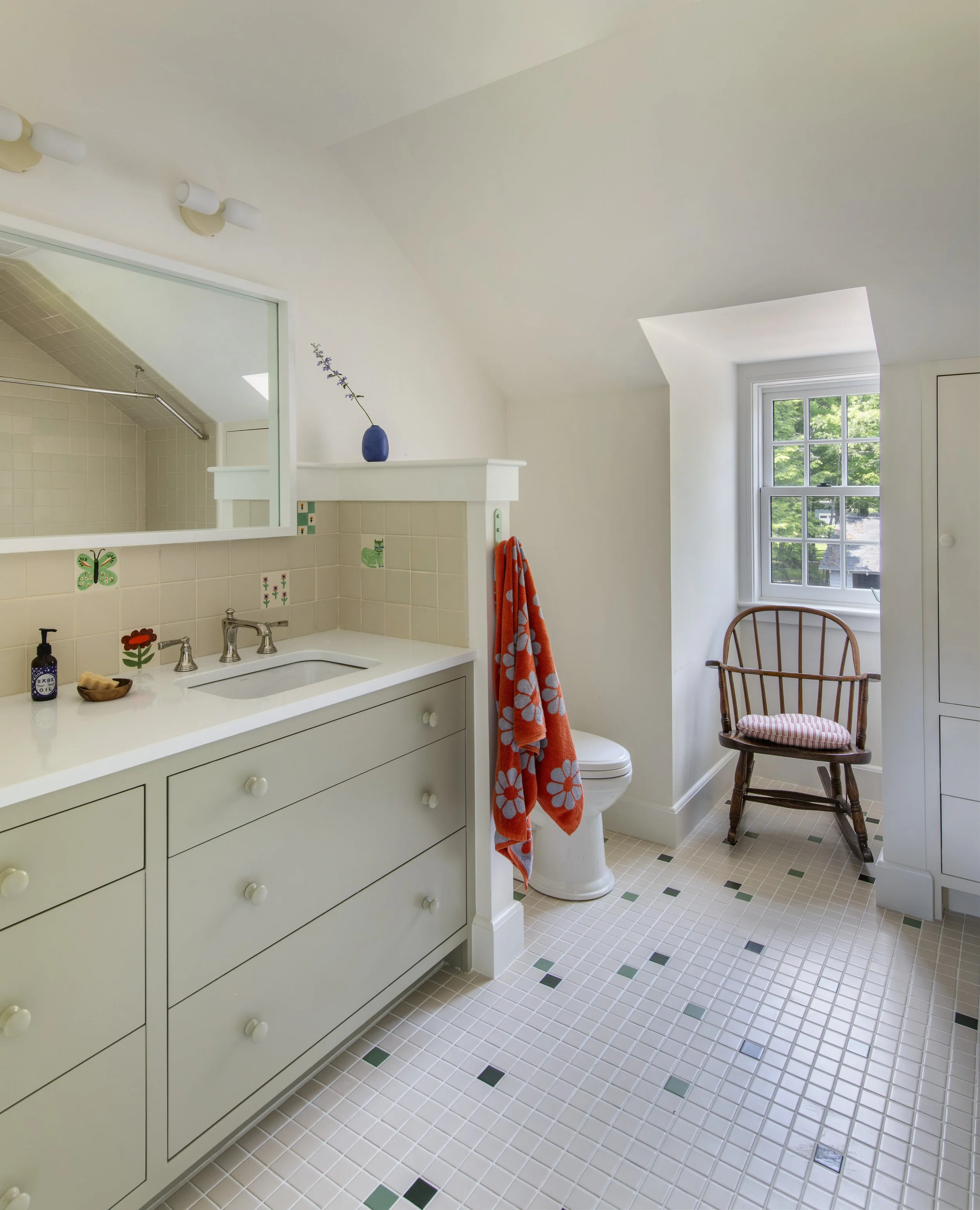 A bathroom with a white vanity, tiled floor, a window with green trees outside, a wooden chair with a striped cushion, and a toilet with a leaning orange towel. A small blue vase with a flower is on a shelf above the toilet. The mirror above the vani
