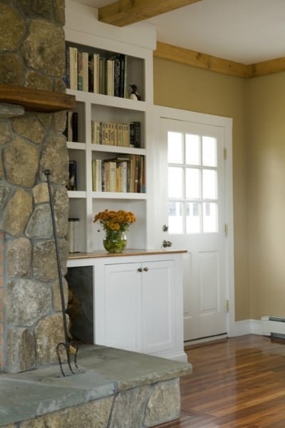 Interior of a cozy room with a stone fireplace, a built-in white bookshelf filled with books, a glass jar of orange flowers on a white cabinet, and a door with glass pane panels leading outside, with hardwood floors and a wooden ceiling beam.