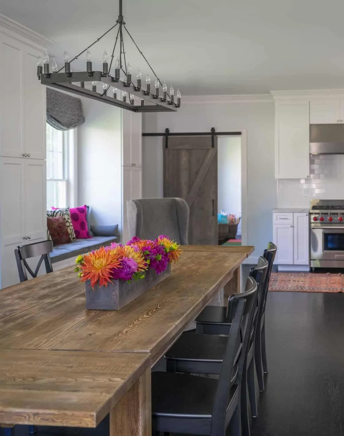 A dining room with a wooden table, black chairs, and a centerpiece of colorful flowers. A chandelier with candle-like bulbs hangs above the table. In the background, a window with a gray valance, a gray armchair, a barn door on a sliding rail, and a 