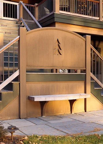 Outdoor wooden privacy screen with a bench, featuring a tree design cutout, located on a patio with stairs and a deck in the background.