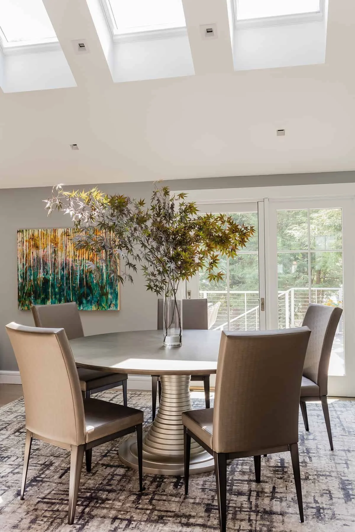 A dining room with a round table, six beige upholstered chairs, a vase with a large green plant, an abstract painting on the wall, large windows with a balcony outside, and a patterned rug.