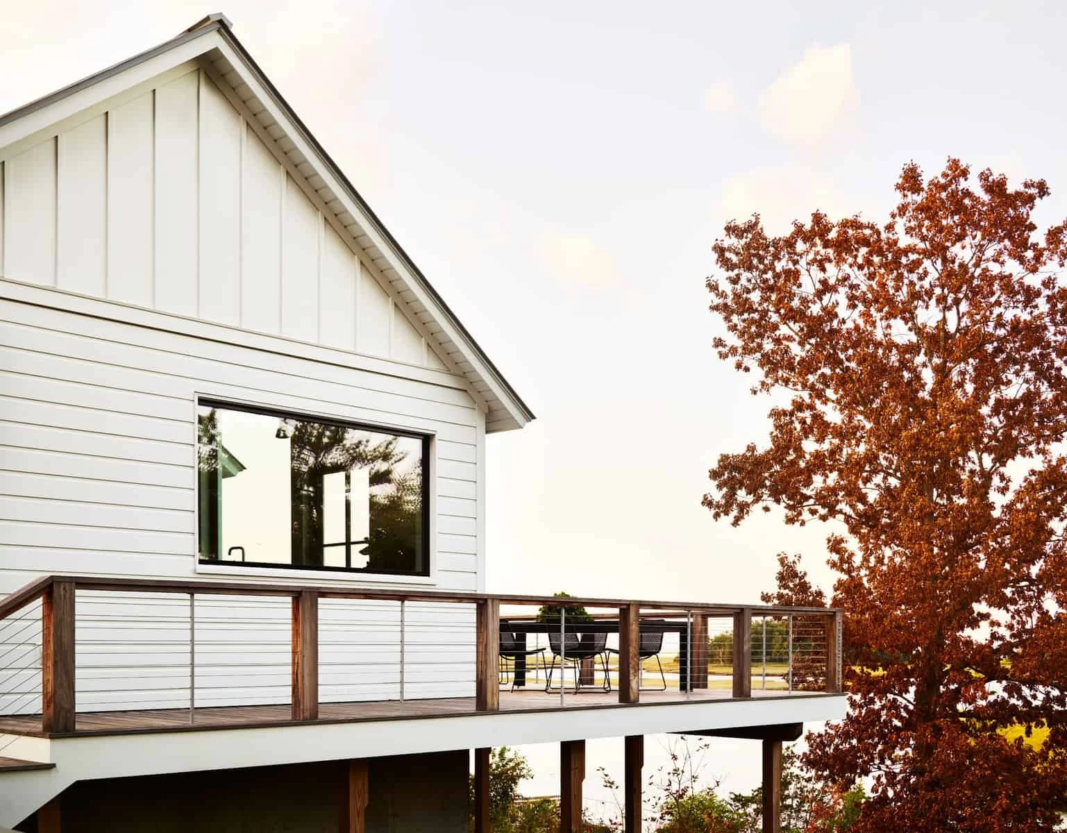 Modern white house with large window and wooden balcony railing, autumn tree with orange leaves, outdoor table and chairs, cloudy sky.