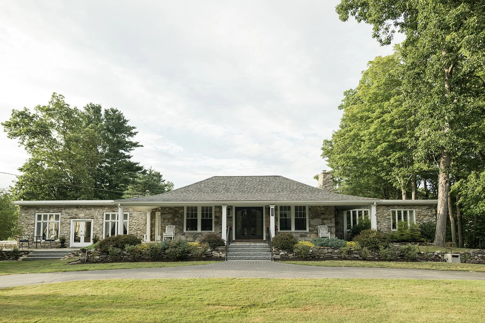A single-story house with stone walls, surrounded by trees and a well-maintained lawn, featuring a central entryway with steps and outdoor seating on the porch.