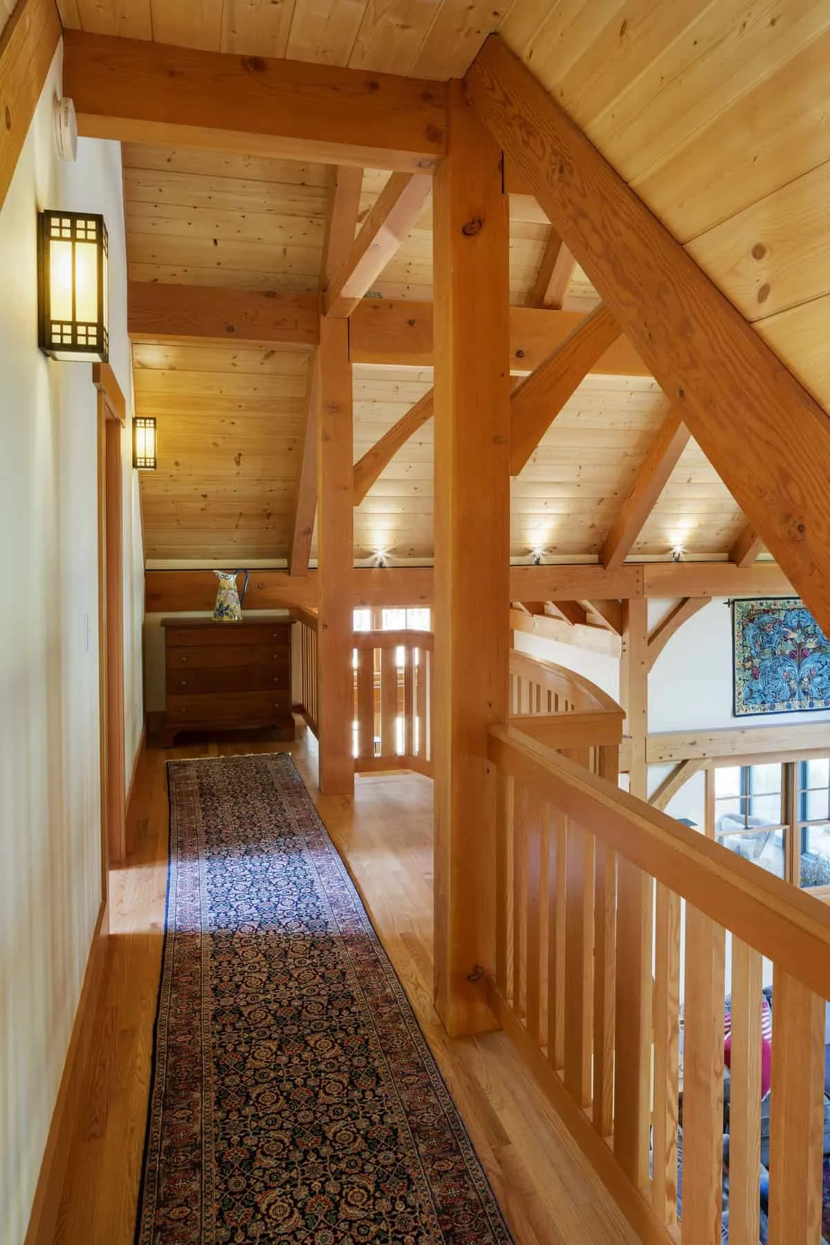Interior view of a wooden home hallway with a staircase, a runner rug, wall-mounted lights, and a piece of colorful artwork on the wall.