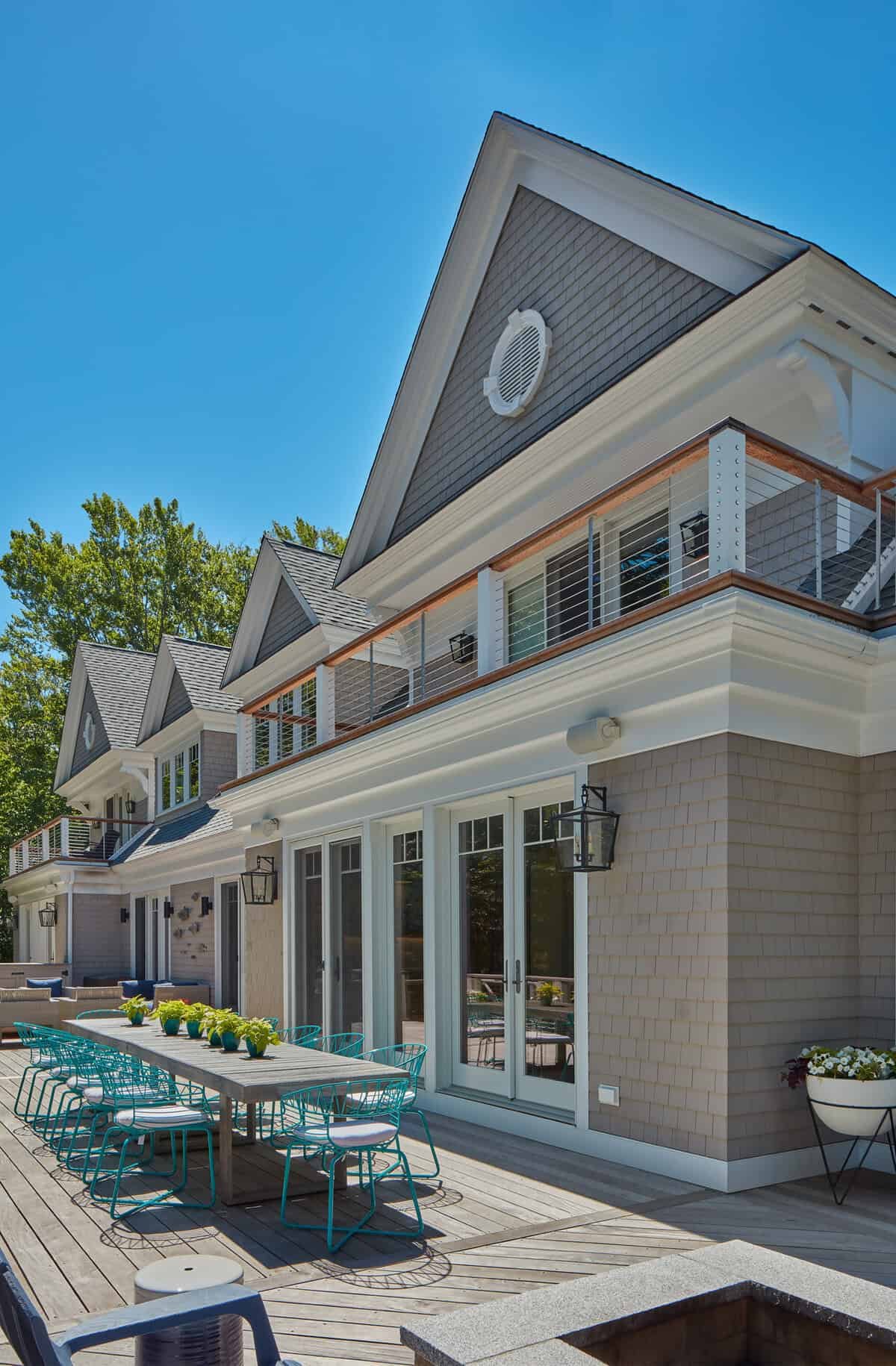 Modern house with a wooden deck outdoor dining area, green potted plants, glass doors, brick walls, and multiple gable roofs under a clear blue sky.