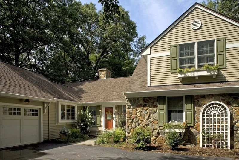 Exterior of a two-story house with stone and siding facade, green shutters, and a garage, surrounded by trees and a small garden.
