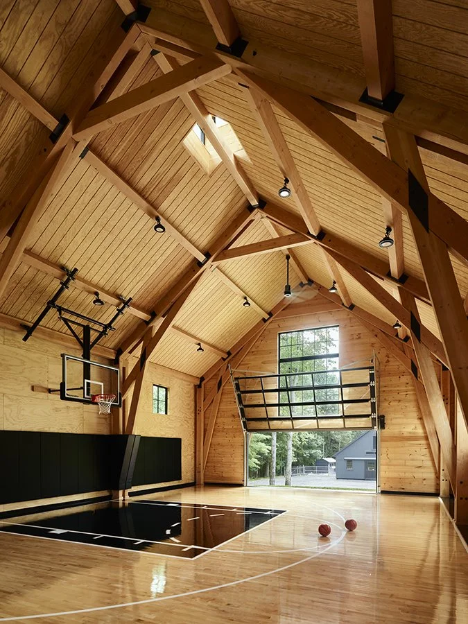 Interior view of a wooden sports barn with a basketball hoop, polished wooden court, and two basketballs on the floor. Large windows show trees outside.