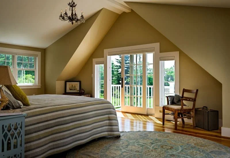 A bright bedroom with beige walls and slanted ceilings, featuring a large bed with a striped bedspread, a side table with a lamp, a wooden chair, and glass doors leading to a small balcony overlooking greenery.