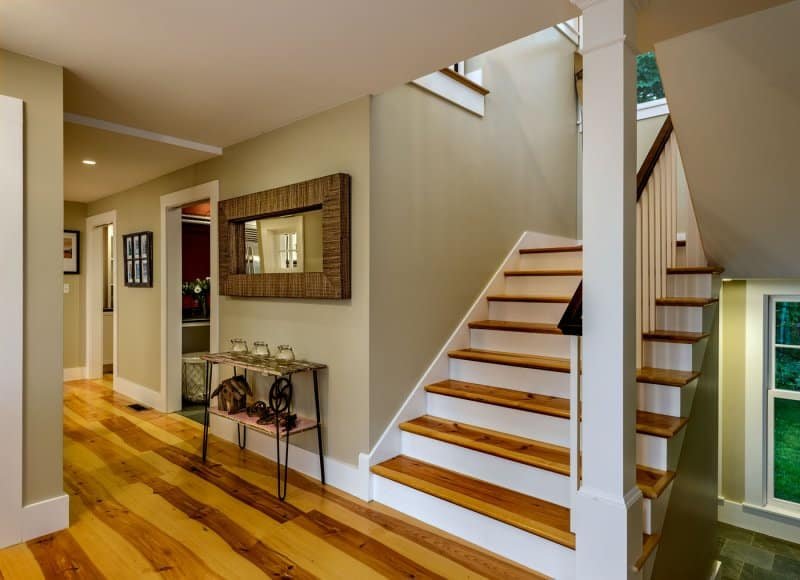 Interior view of a modern home featuring a wooden staircase with white risers and beige walls; adjacent hallway has hardwood flooring, framed artwork, and a decorative console table.