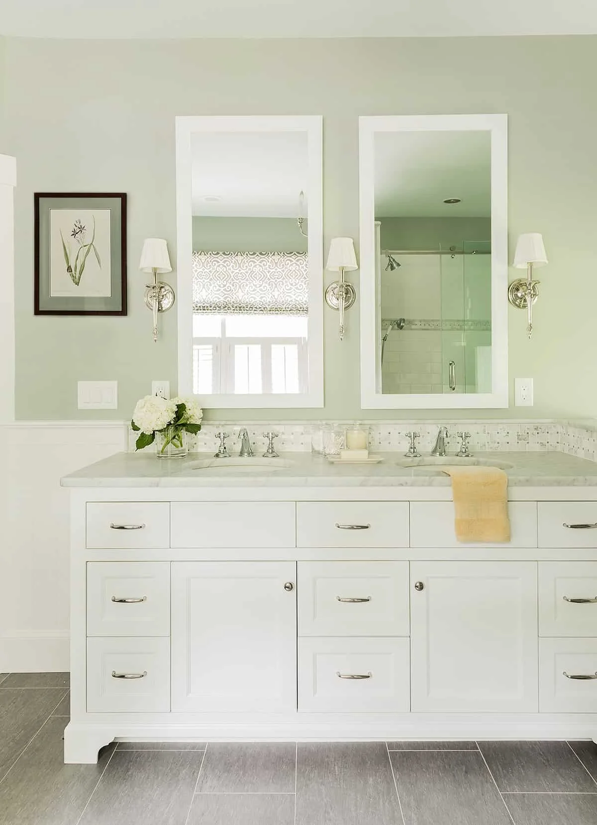 A white bathroom vanity with a marble countertop, adorned with a vase of white flowers, and two mirrors with wall-mounted lamps. A yellow towel hangs over the right side of the vanity, and the background features a light green wall with a framed bota