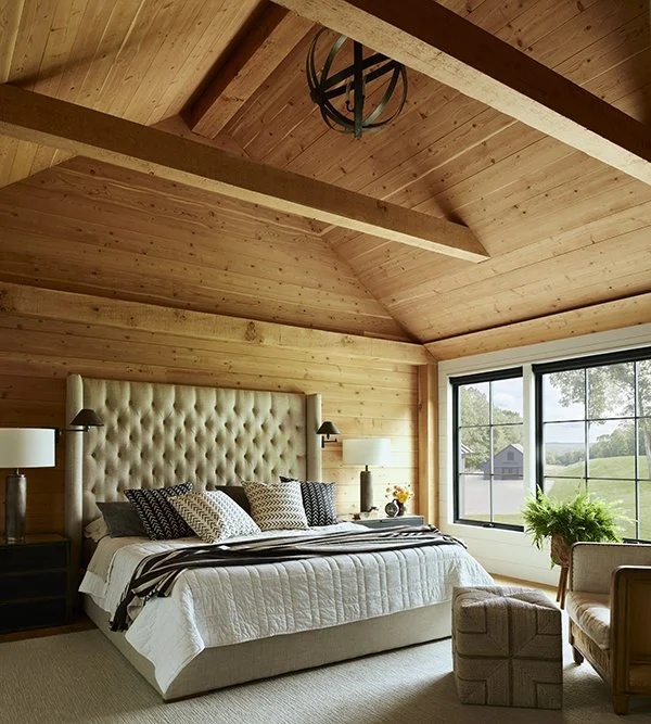 Bedroom with wood-paneled vaulted ceiling, large window showing outdoor landscape, beige upholstered bed with pillows, side tables with lamps, armchair, and a potted plant.
