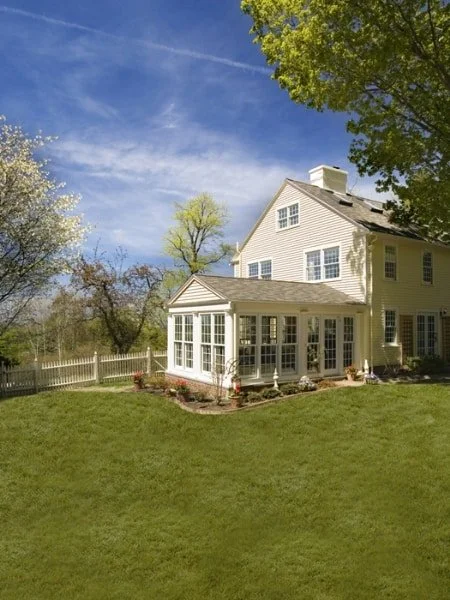 A large, white, two-story house with multiple windows and a sunroom, surrounded by green grass, trees, and a clear blue sky.