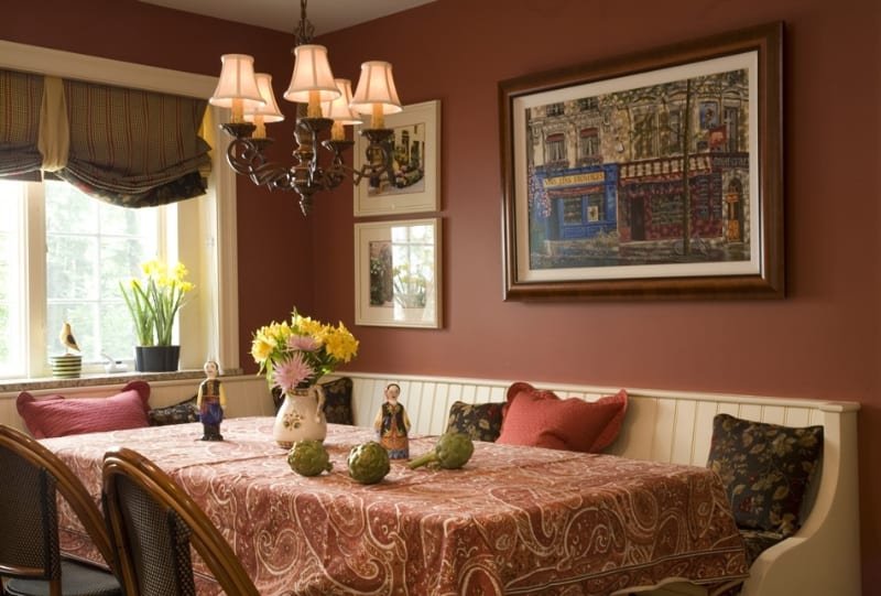 A dining room with a floral tablecloth, decorative vases, and small figurines. There is a window with yellow flowers, framed artwork on the walls, and a chandelier hanging above the table.