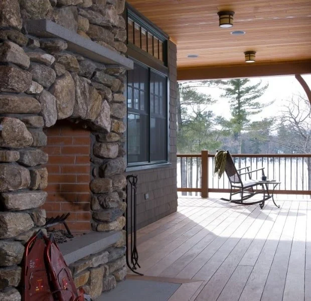 Covered porch with wooden flooring and a wooden rocking chair, overlooking trees and a snow-covered landscape, with a stone fireplace on the left.