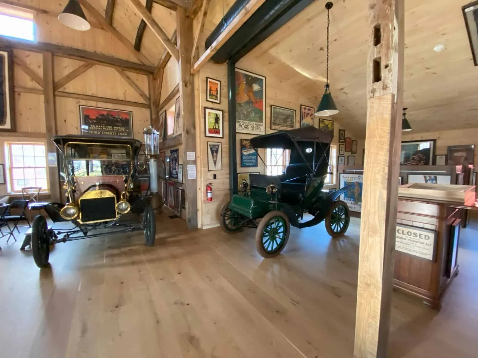 Interior of a museum with vintage cars displayed on a wooden floor, wood-paneled walls, and framed posters and pictures hanging inside.