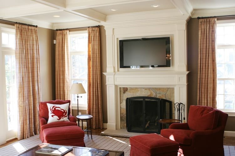 Living room with fireplace, flat-screen TV above mantle, two red armchairs, side table, floor lamp, and large windows with plaid curtains.