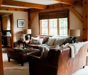 Cozy living room with wooden beams, large windows, a brown leather sofa with patterned cushions, and a matching leather ottoman.