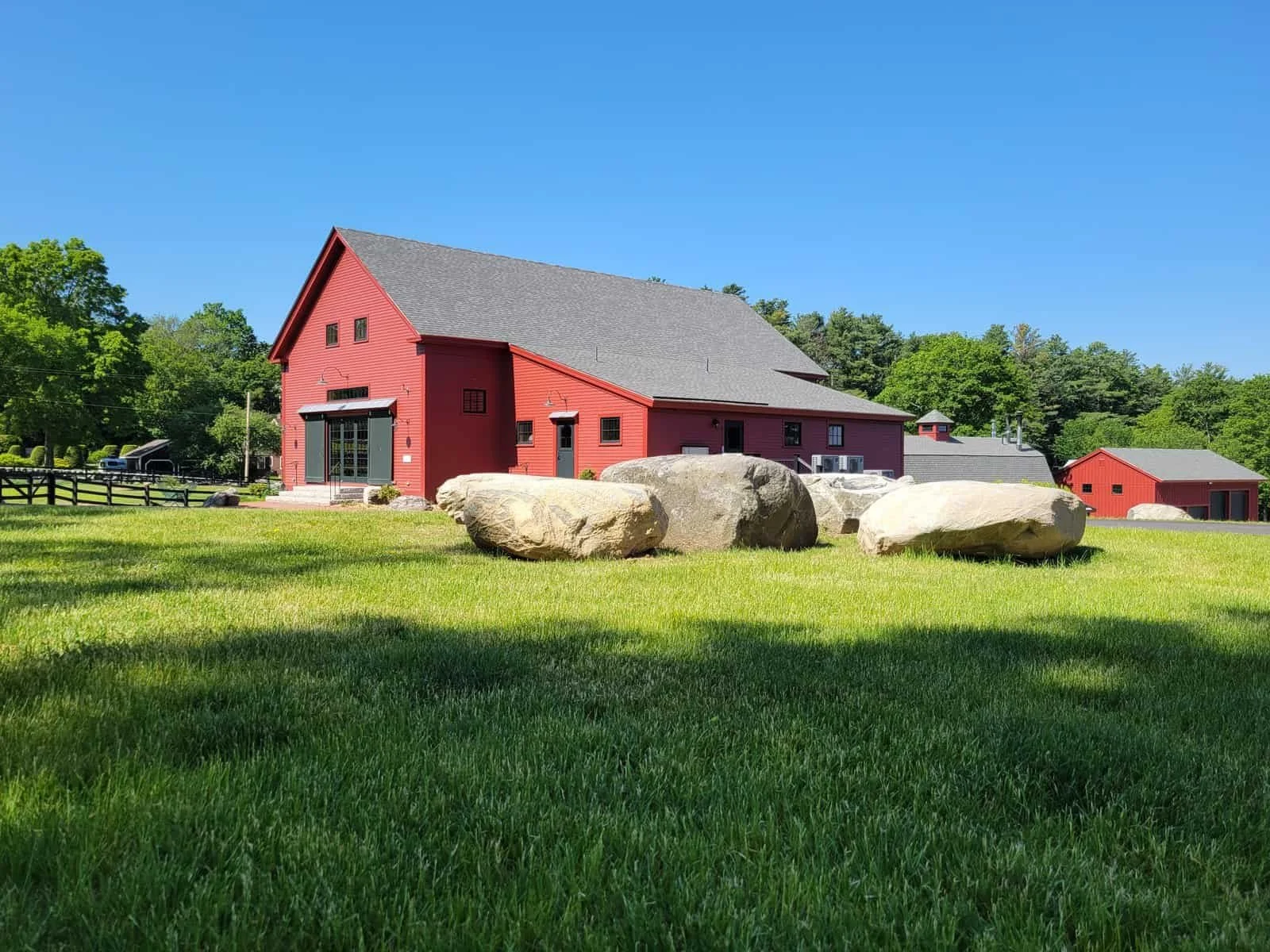 A red barn with a gray roof on a grassy field, large boulders in the foreground, trees in the background, under a clear blue sky.
