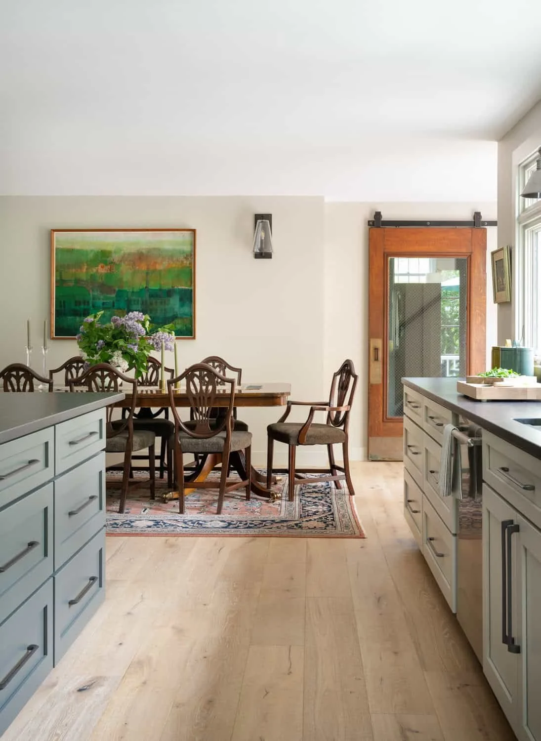 Interior view of a kitchen and dining area with light hardwood floors, white cabinets, a wooden dining table with six chairs, a colorful painting on the wall, a sliding glass door, and a window with sunlight streaming in.