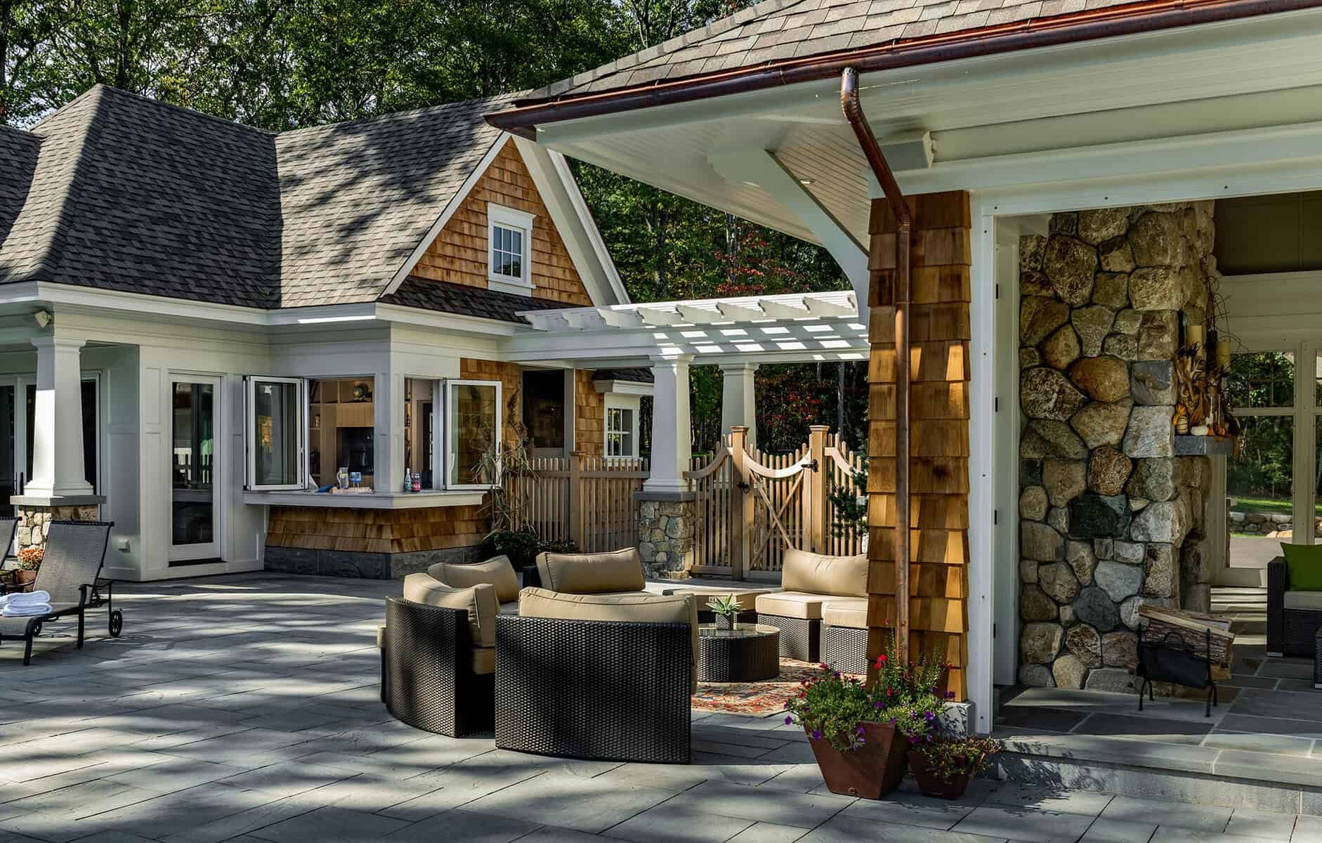 A spacious outdoor patio area with patio furniture, including chairs and a table with an umbrella. The house features a mix of white siding, natural wood shingles, and stone accents, with trees visible in the background.